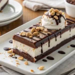Close-up of a layered hot fudge ice cream bar dessert slice with visible fudge, whipped topping, and chopped peanuts on a white plate